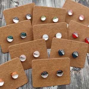 May include: Assortment of small, round, two-toned earrings displayed on brown cards. The earrings are half silver and half a variety of colors, including green, red, black, and blue.