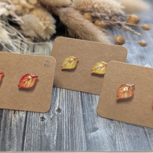 Puede incluir: Tres pares de pendientes de bot&oacute;n con forma de hoja sobre tarjetas marrones. Los pendientes presentan colores oto&ntilde;ales de rojo, naranja y amarillo. El fondo incluye flores secas y una superficie de madera.