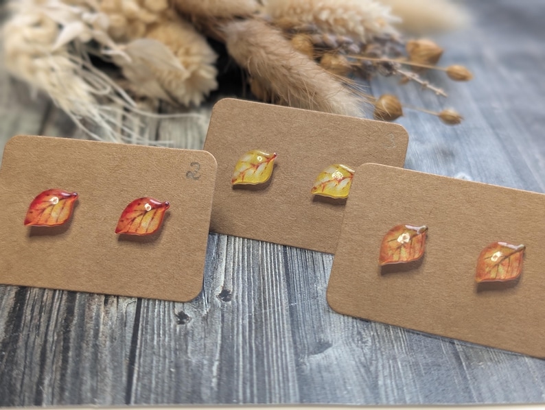 Puede incluir: Tres pares de pendientes de bot&oacute;n con forma de hoja sobre tarjetas marrones. Los pendientes presentan colores oto&ntilde;ales de rojo, naranja y amarillo. El fondo incluye flores secas y una superficie de madera.