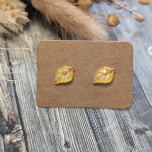 Puede incluir: Un par de pendientes de bot&oacute;n con forma de hoja. Cada pendiente presenta un dise&ntilde;o de hoja amarilla con vetas rojas. Los pendientes se muestran en una tarjeta marr&oacute;n, sobre un fondo de madera con acentos florales secos.
