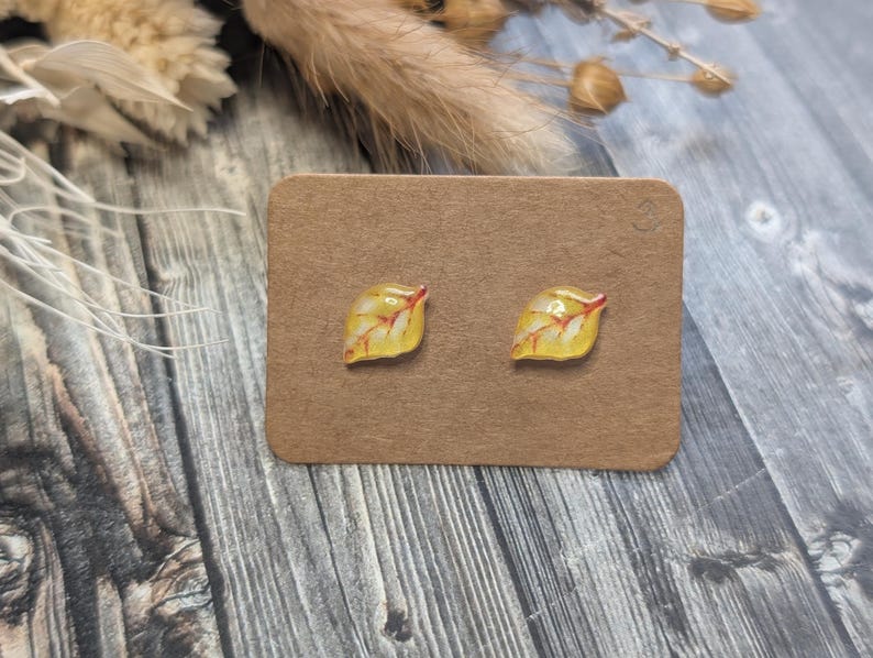 Puede incluir: Un par de pendientes de bot&oacute;n con forma de hoja. Cada pendiente presenta un dise&ntilde;o de hoja amarilla con vetas rojas. Los pendientes se muestran en una tarjeta marr&oacute;n, sobre un fondo de madera con acentos florales secos.