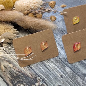 Puede incluir: Pendientes con dise&ntilde;os de hojas en tonos amarillos, naranjas y marrones se exhiben en tarjetas marrones. Los pendientes se colocan sobre un fondo de madera r&uacute;stica, con flores secas y follaje en la esquina superior izquierda.