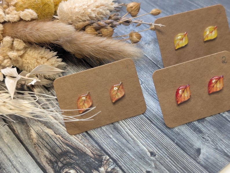 Puede incluir: Pendientes con dise&ntilde;os de hojas en tonos amarillos, naranjas y marrones se exhiben en tarjetas marrones. Los pendientes se colocan sobre un fondo de madera r&uacute;stica, con flores secas y follaje en la esquina superior izquierda.