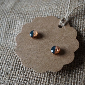 May include: A pair of small, round earrings with a black and copper design. The earrings are displayed on a brown card with scalloped edges.