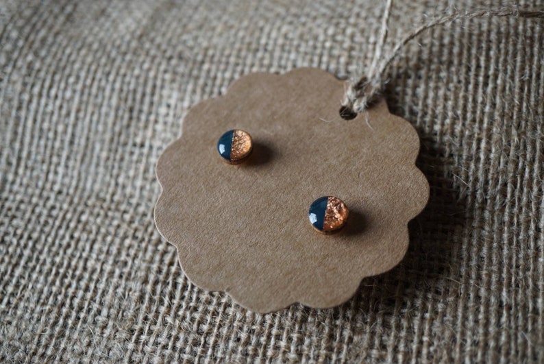 May include: A pair of small, round earrings with a black and copper design. The earrings are displayed on a brown card with scalloped edges.