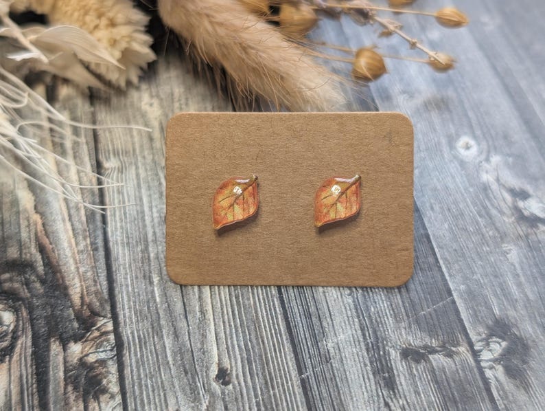 Puede incluir: Un par de pendientes de bot&oacute;n con forma de hoja. Los pendientes son de color marr&oacute;n claro con detalles en naranja y amarillo, y una peque&ntilde;a gema transparente en el centro. Se presentan sobre una tarjeta marr&oacute;n.