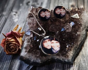 Puede incluir: Un conjunto de collar y pendientes de plata con colgantes redondos de fotos que muestran a una pareja. Los colgantes están colocados sobre un fondo de cristal gris oscuro.