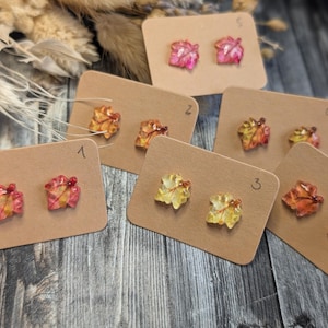 Puede incluir: Un conjunto de pendientes de botón con forma de hoja en varios colores, incluyendo rosa, amarillo y naranja, expuestos en pequeñas tarjetas marrones. Los pendientes están dispuestos sobre una superficie de madera, mostrando detalles.