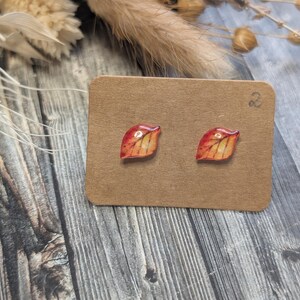 Puede incluir: Pendientes de bot&oacute;n con forma de hoja, pintados en tonos rojos, naranjas y amarillos, que recuerdan al follaje oto&ntilde;al. Se exhiben sobre una peque&ntilde;a tarjeta de cart&oacute;n marr&oacute;n. El fondo es de madera.