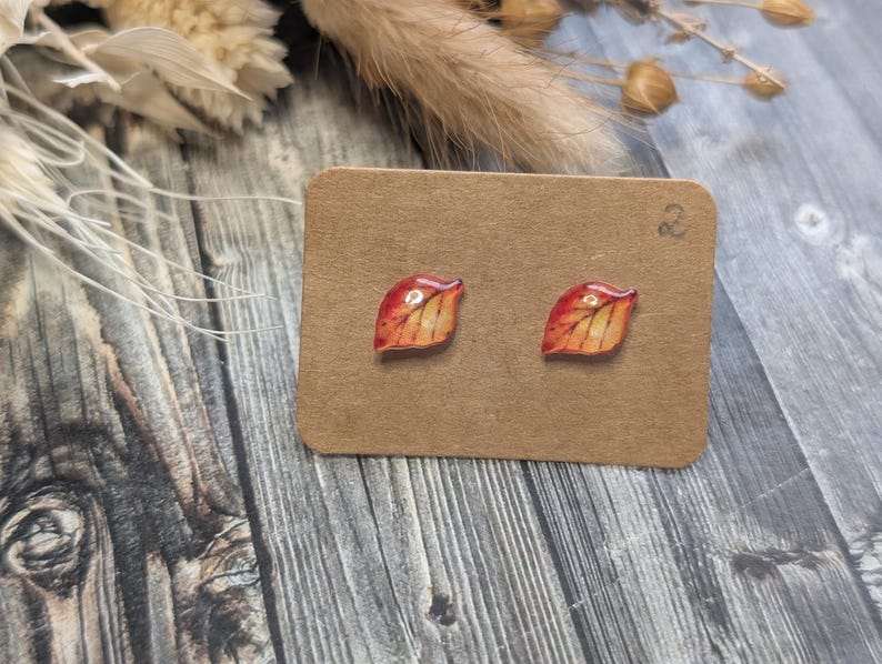 Puede incluir: Pendientes de bot&oacute;n con forma de hoja, pintados en tonos rojos, naranjas y amarillos, que recuerdan al follaje oto&ntilde;al. Se exhiben sobre una peque&ntilde;a tarjeta de cart&oacute;n marr&oacute;n. El fondo es de madera.
