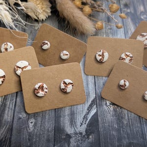 May include: Stud earrings with a white base and copper-colored veining are displayed on brown cardboard cards. The earrings are round and come in various sizes. The background features dried flowers and a wooden surface.