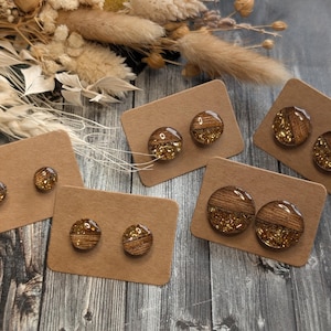 Peut inclure: Plusieurs paires de boucles d'oreilles en bois avec des accents de paillettes dorées sont présentées sur des cartons bruns. Les boucles d'oreilles sont rondes, avec un motif de grain de bois et des paillettes incrustées. Des fleurs séchées sont en arrière-plan.