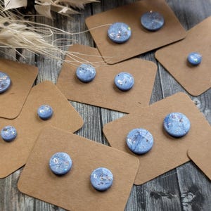 May include: A collection of blue and silver stud earrings displayed on small, brown cardboard tags. The earrings have a marbled appearance with embedded glitter. The background is a dark wood grain surface.