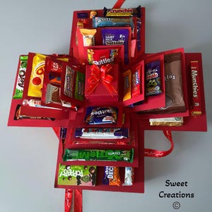 May include: A red explosion box, packed with an assortment of sweets and chocolates. The box, adorned with a red ribbon, features popular brands such as Skittles, Crunchie, and Dairy Milk. The box is labelled "Sweet Creations".