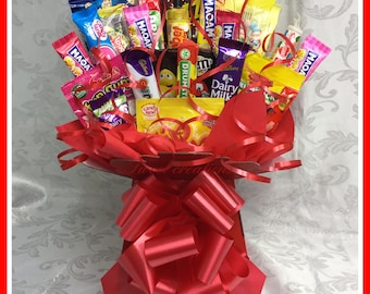 May include: A red gift box filled with a variety of colorful candy bars and chocolate. The box is tied with a red ribbon and bow. The candy includes brands like Dairy Milk, Twix, and M&Ms.
