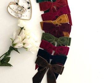 May include: An assortment of velvet hair bows in various colors, including green, burgundy, and brown, are arranged on a white surface. The bows have a classic tied design. A heart-shaped dish with jewelry and a bouquet of white flowers are also present.