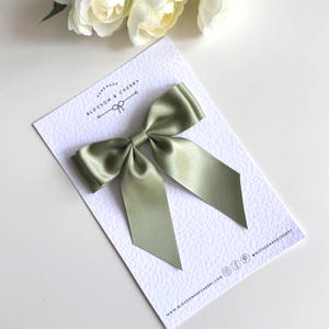 Può includere: Un fiocco di raso verde salvia su una carta bianca strutturata. Il fiocco è centrato sulla carta, con lunghe code a punta che si estendono dal nodo. La carta ha il testo "HANDMADE BLOSSOM & CHERRY" e le icone dei social media.