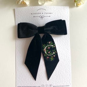 May include: Black velvet bow with a white letter C embroidered with pink and green floral details. The bow is handmade by Blossom & Cherry. The text "HANDMADE" and "BLOSSOM & CHERRY" are printed on the card. The website address "www.blossomandcherry.com" and social media icons are printed on the bottom of the card.