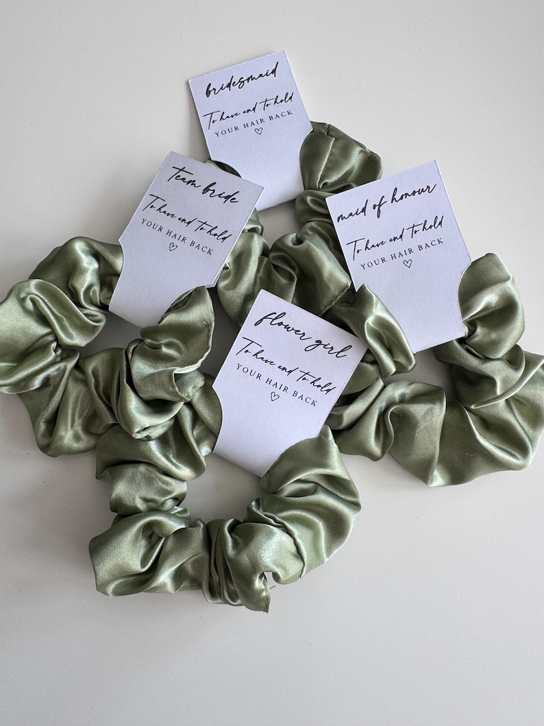 May include: Sage green satin scrunchies with white tags. Each tag has a different title: "bridesmaid", "maid of honour", "team bride", and "flower girl". The tags read "To have and to hold YOUR HAIR BACK" with a heart symbol.