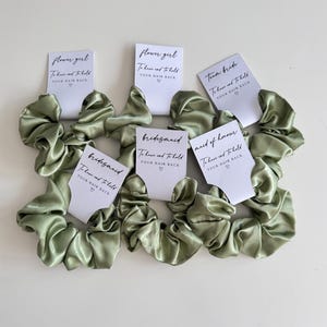 May include: Sage green satin scrunchies with white cards that read "Flower Girl", "Bridesmaid", "Team Bride", and "Maid of Honour". Each card has the text "To have and to hold YOUR HAIR BACK" with a heart symbol.