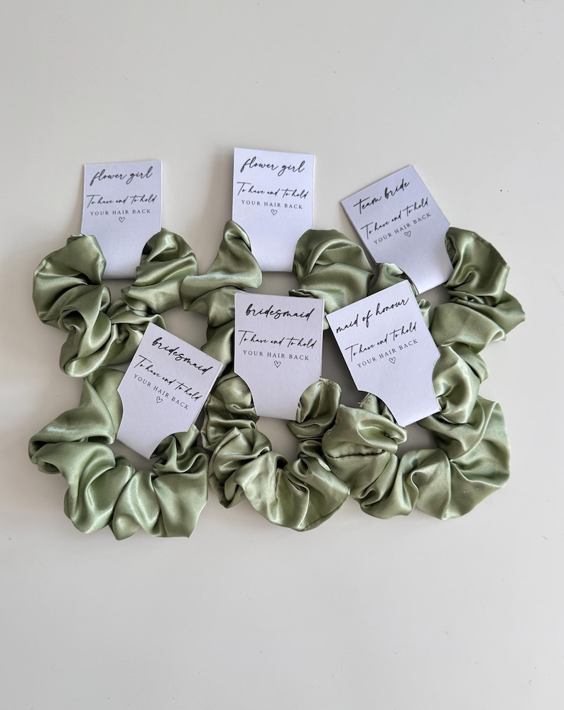May include: Sage green satin scrunchies with white cards that read "Flower Girl", "Bridesmaid", "Team Bride", and "Maid of Honour". Each card has the text "To have and to hold YOUR HAIR BACK" with a heart symbol.