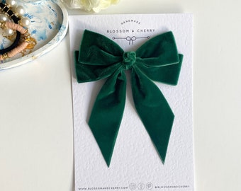 May include: A handmade, emerald green velvet bow on a white card. The bow is centered on the card, with long, pointed tails. The card has the text "BLOSSOM & CHERRY" and social media icons at the bottom.
