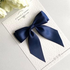 May include: A navy blue satin bow with tails, handmade by Blossom & Cherry. The bow is displayed on a white card with the company's logo and website address.