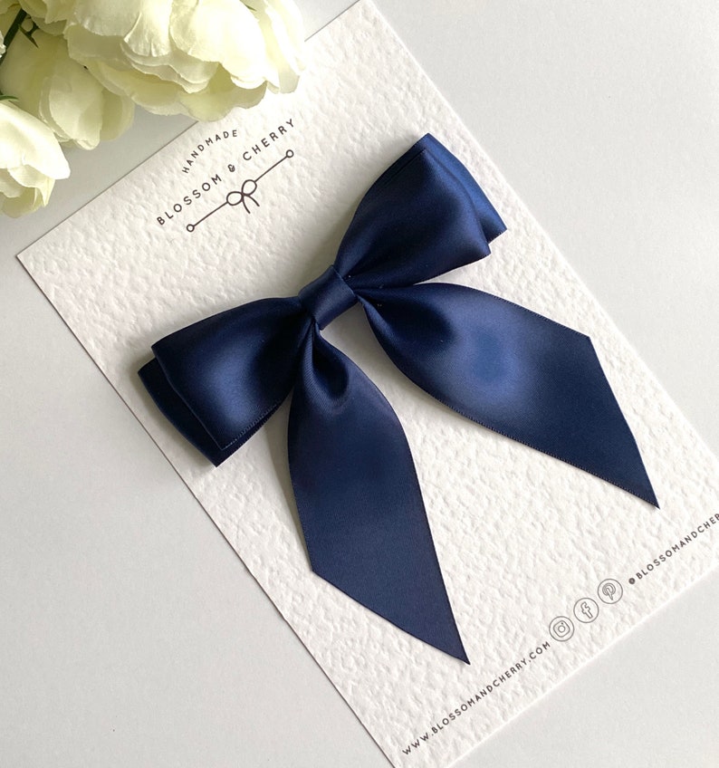 May include: A navy blue satin bow with tails, handmade by Blossom & Cherry. The bow is displayed on a white card with the company's logo and website address.