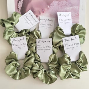 May include: Sage green satin scrunchies with white tags that read "bridesmaid", "maid of honour", "flower girl", and "team bride". Each tag has the text "To have and to hold YOUR HAIR BACK" with a heart symbol. The scrunchies are arranged on a magazine.