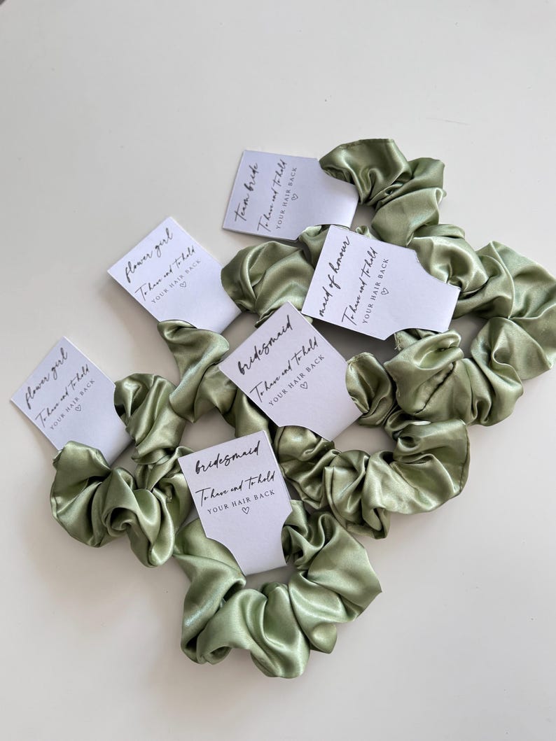 May include: Sage green satin scrunchies with white cards. The cards have black text that reads "flower girl", "Team Bride", "bridesmaid", and "maid of honor". Each card also has the text "To have and to hold YOUR HAIR BACK".