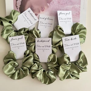 May include: Olive green satin scrunchies with white cards that read "bridesmaid", "maid of honor", "flower girl", and "team bride". Each card says "To have and to hold YOUR HAIR BACK" with a heart symbol. The scrunchies are on a light background.