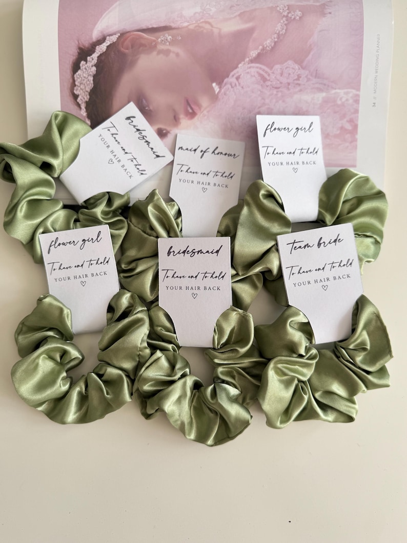 May include: Olive green satin scrunchies with white cards that read "bridesmaid", "maid of honor", "flower girl", and "team bride". Each card says "To have and to hold YOUR HAIR BACK" with a heart symbol. The scrunchies are on a light background.