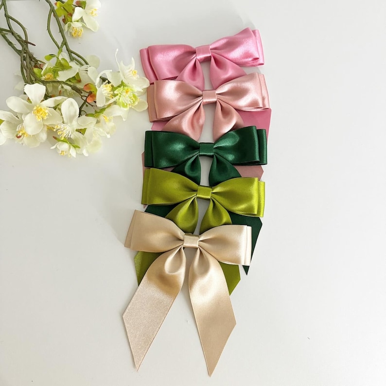 May include: Five satin ribbon bows in different colors. The bows are arranged vertically, with the largest bow at the bottom and the smallest bow at the top. The colors of the bows are pink, green, and beige.