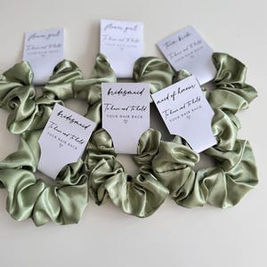 May include: Sage green satin scrunchies with white cards that read "bridesmaid", "maid of honour", and "flower girl". Each card has the text "To have and to hold YOUR HAIR BACK" in black script.