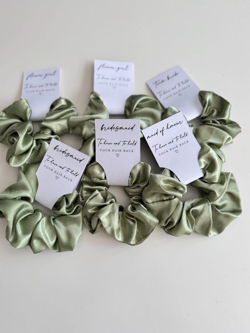 May include: Sage green satin scrunchies with white cards that read "bridesmaid", "maid of honour", and "flower girl". Each card has the text "To have and to hold YOUR HAIR BACK" in black script.
