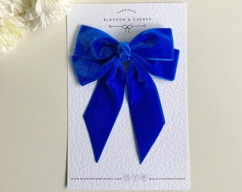 May include: A bright blue velvet bow with a center knot. The bow is handmade and is displayed on a white card with the text "Handmade Blossom & Cherry" and the website address "www.blossomandcherry.com".