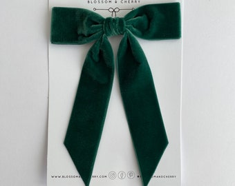 May include: A dark green velvet bow with long tails. The bow is handmade and is tied with a knot in the center. The bow is on a white background with the text "HANDMADE BLOSSOM & CHERRY" above it.