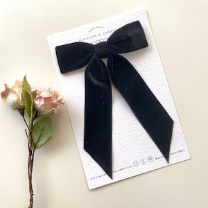 Può includere: Fiocco in velluto nero con code lunghe, fatto a mano da Blossom & Cherry. Il fiocco è esposto su una carta bianca con il nome dell'azienda e l'indirizzo del suo sito web.