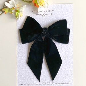 Pode incluir: Um grande la&ccedil;o de veludo preto com caudas longas. O la&ccedil;o &eacute; amarrado em um n&oacute; no centro e as caudas s&atilde;o pontiagudas nas pontas. O la&ccedil;o est&aacute; sobre um fundo branco com o texto "Blossom & Cherry" impresso nele.