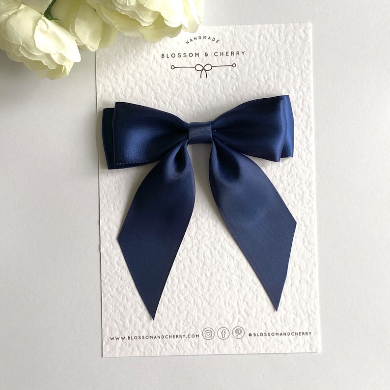 May include: A navy blue satin bow with pointed ends. The bow is handmade and is displayed on a white card with the text "Handmade Blossom & Cherry" and the website address "www.blossomandcherry.com".
