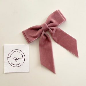 May include: A dusty rose velvet bow with long tails. The bow is handmade and has a circular logo with the text "Blossom & Cherry Handmade" in black.