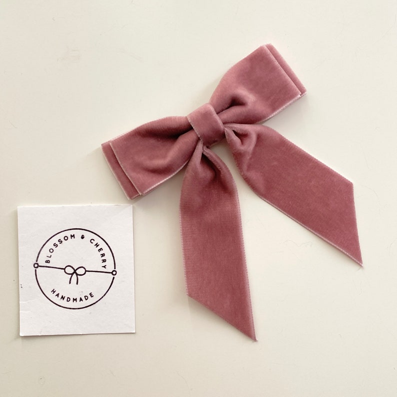 May include: A dusty rose velvet bow with long tails. The bow is handmade and has a circular logo with the text "Blossom & Cherry Handmade" in black.