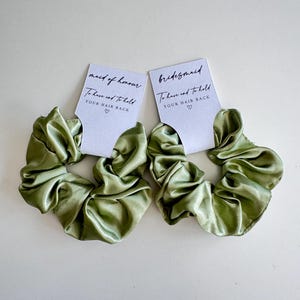 May include: Two olive green satin scrunchies with white tags. The tags read "maid of honour" and "bridesmaid" with the text "To have and to hold YOUR HAIR BACK" and a heart symbol. The scrunchies are on a white background.