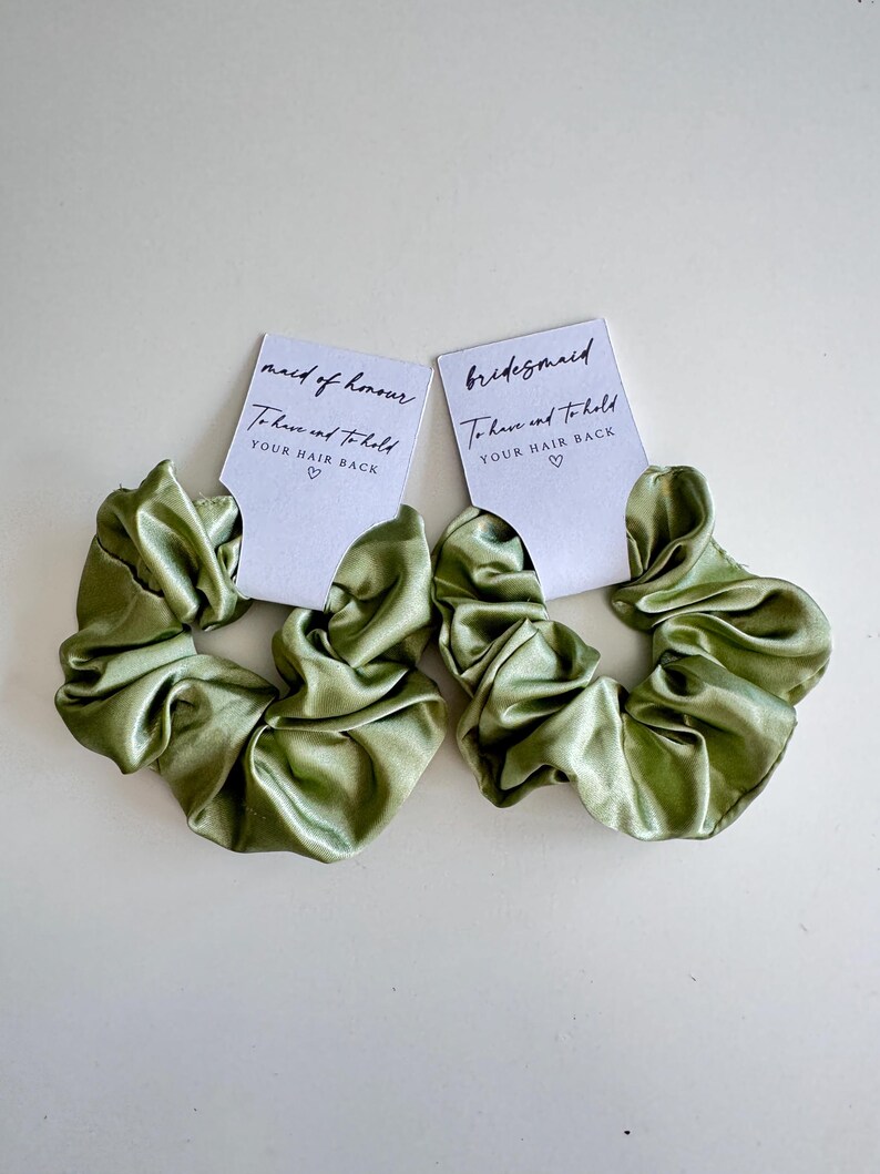 May include: Two olive green satin scrunchies with white tags. The tags read "maid of honour" and "bridesmaid" with the text "To have and to hold YOUR HAIR BACK" and a heart symbol. The scrunchies are on a white background.