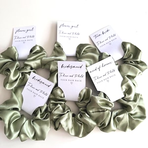 May include: Sage green satin scrunchies with white cards. The cards read "flower girl", "bridesmaid", "maid of honour", and "team bride". Each card has the text "To have and to hold YOUR HAIR BACK" with a heart symbol.