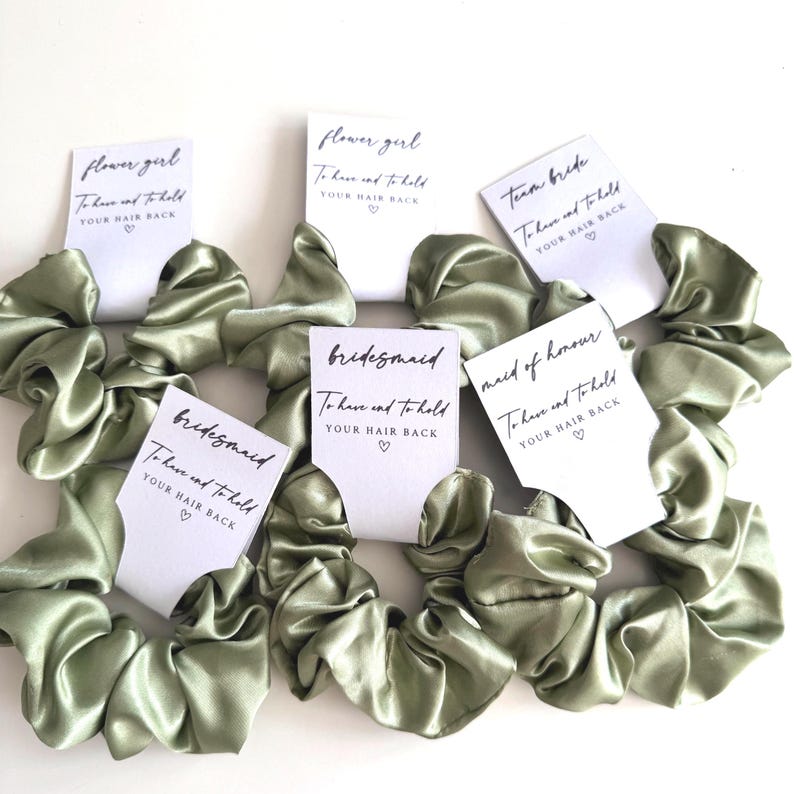 May include: Sage green satin scrunchies with white cards. The cards read "flower girl", "bridesmaid", "maid of honour", and "team bride". Each card has the text "To have and to hold YOUR HAIR BACK" with a heart symbol.