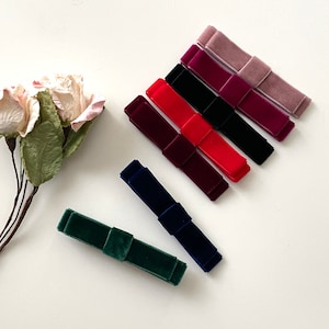 May include: Assortment of six velvet hair bows in various colors, including pink, red, burgundy, black, navy blue, and green. The bows are arranged in a row on a white surface.