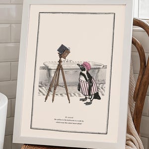 May include: A framed print with a humorous illustration of a penguin in a striped coat and pink hat, using an old camera on a tripod. The penguin is near a bathtub. The print is displayed on a wooden chair, in a bathroom setting.