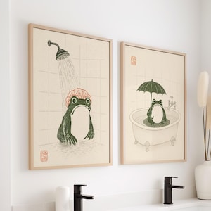 Könnte beinhalten: Zwei gerahmte Kunstdrucke mit Froschillustrationen. Einer zeigt einen Frosch unter der Dusche, der andere in einer Badewanne mit einem Regenschirm. Die Drucke haben einen beigefarbenen Hintergrund mit einem Rastermuster und sind in hellen Holzrahmen.