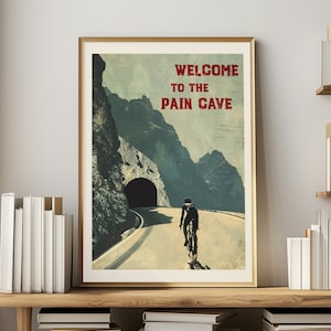 Può includere: Un poster in stile vintage con un ciclista che sale su una strada di montagna verso un tunnel. Il poster ha il testo "WELCOME TO THE PAIN CAVE" in rosso.